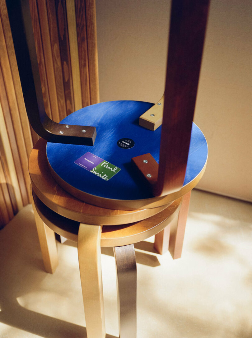 Two stacked Artek wooden stools with a blue seat and stickers facing upwards under warm lighting. The visible stool has four attached legs and labels indicating design details, reflecting Paul Smith's influence.