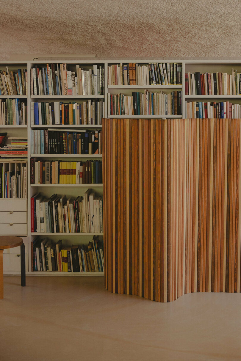 Bookshelf filled with various books, partially obscured by a Paul Smith wooden room divider with vertical slats. A chair from the Artek collection is partially visible to the left. The scene appears in a well-lit room.