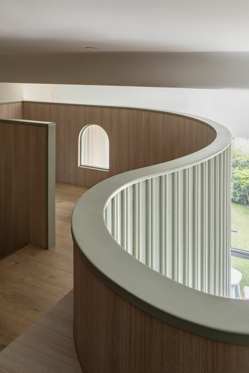 Curved interior balcony with a light green railing and slatted design. Wooden flooring and wall, with an arched window in the background overlooking a garden.