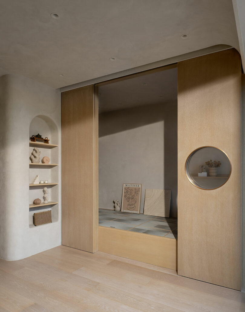 Minimalist room with wooden sliding doors, round cutout in the right door, built-in shelves to the left displaying decor items, and a low platform inside holding art prints. Neutral color palette.