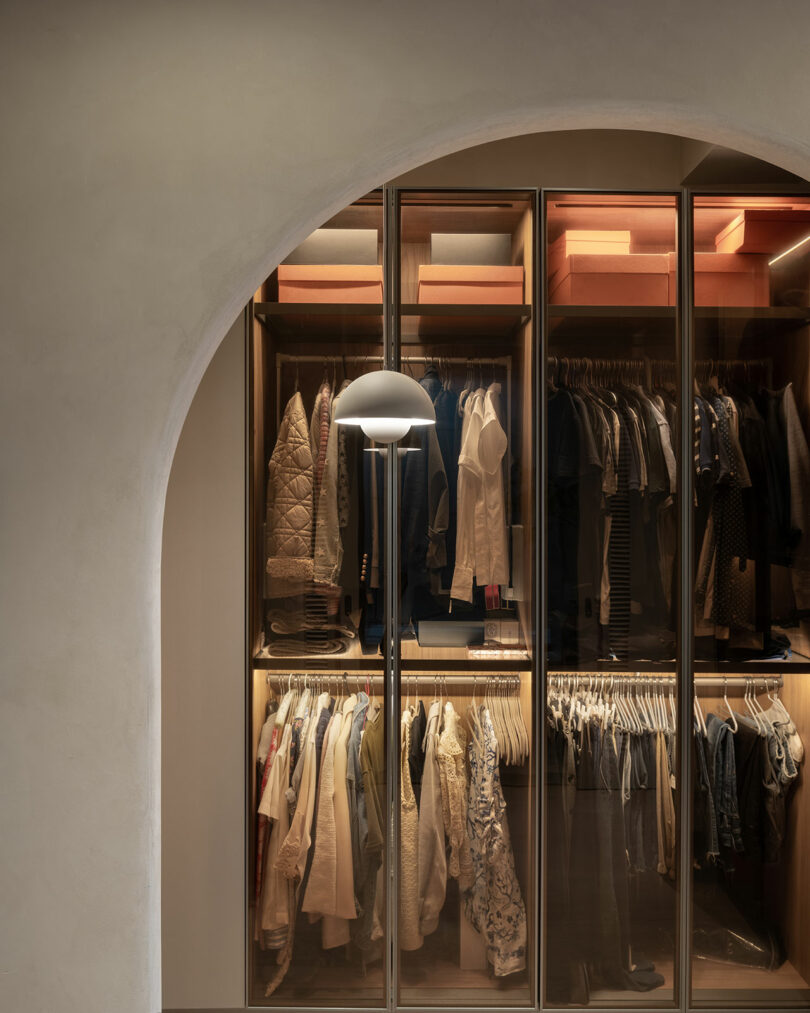 A neatly organized walk-in closet with clothes and shoes on shelves and hangers, illuminated by a floor lamp. An arched doorway frames the entrance.