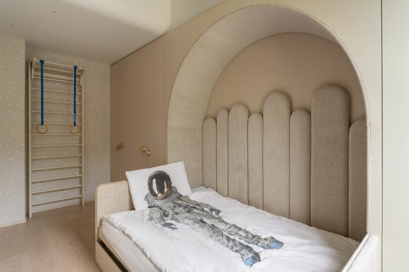 A child's bedroom with a neutral color scheme features a bed with an astronaut-themed pillow and a soft, curved headboard design. The room also includes a climbing wall and rings for physical activity.