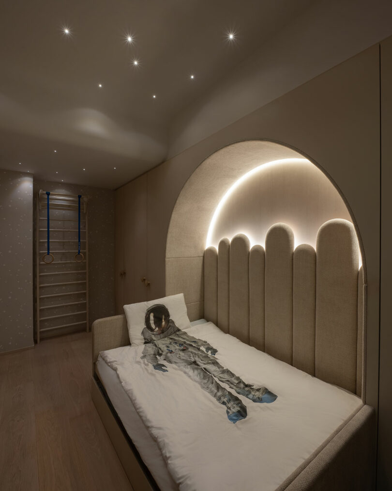 A child's bedroom featuring a bed with an astronaut-themed bedspread and a starry ceiling. The bed's headboard is illuminated with soft, curved lights. Gymnastics rings and ladder are on the wall.