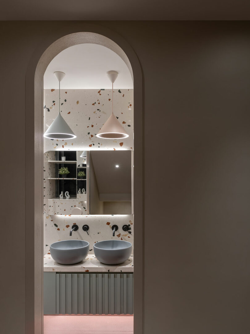 A modern bathroom viewed through an arched doorway, featuring two round sinks, wall-mounted faucets, pendant lights, and a terrazzo backsplash.