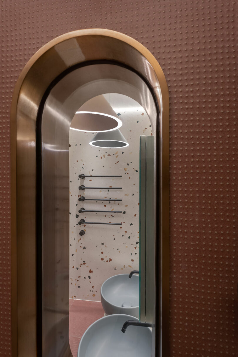 View through an arched mirror showing a modern bathroom with terrazzo wall, two sinks, and black towel racks.