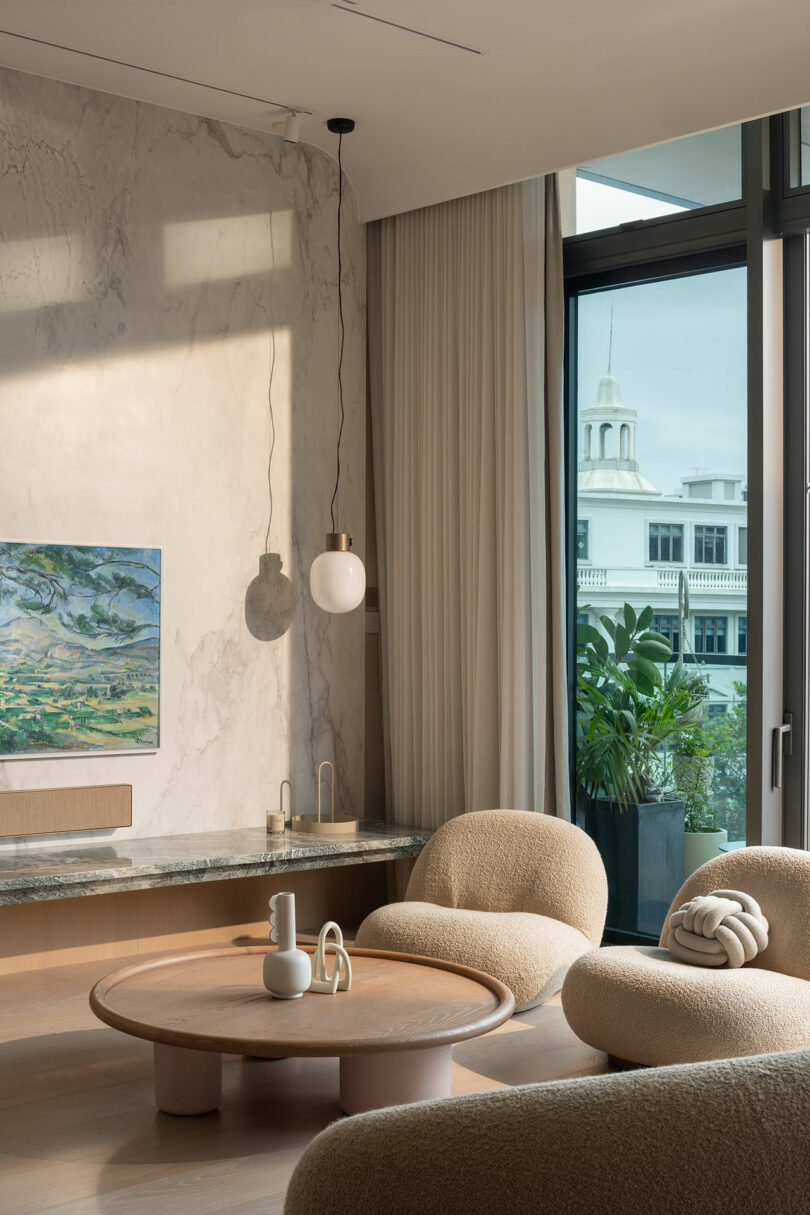 Modern living room with plush beige chairs, round wooden coffee table, art on marble wall, contemporary pendant light, large windows with sheer curtains, and cityscape view featuring a white building.