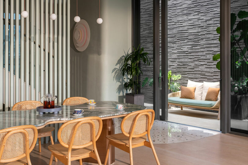 Modern dining area with a marble table, rattan chairs, hanging lights, and a wall-mounted decorative piece. A glass door opens to a patio with a cushioned bench, potted plants, and a textured stone wall.