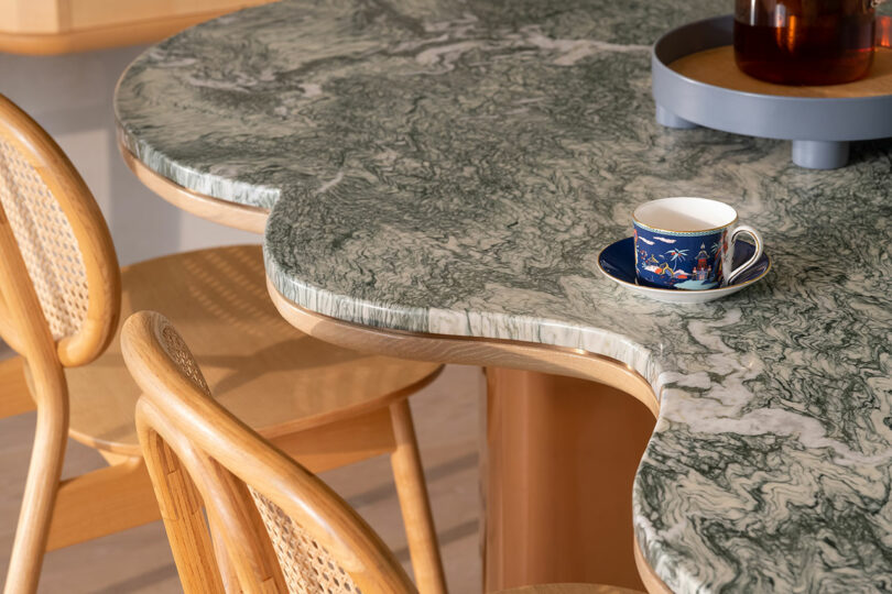 A small teacup sits on a green marble table with curved edges, beside a wooden chair with a woven back.