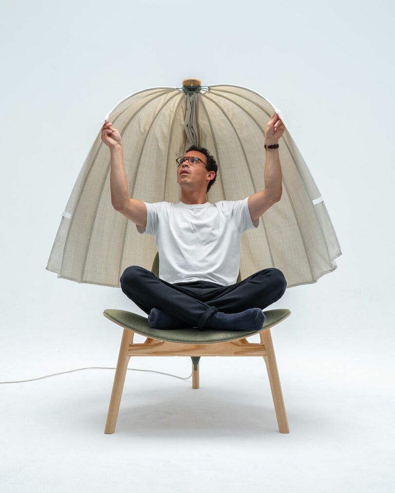 A person seated cross-legged on a Shanti Chair under a dome-like cover, adjusting the edges. The cover extends from the top of the chair to above the person's head.