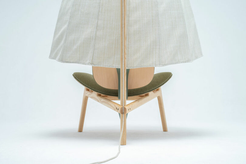 A wooden Shanti Chair with a green seat cushion is positioned under a large, light-colored fabric lampshade hanging from above, with an electrical cord extending from it.