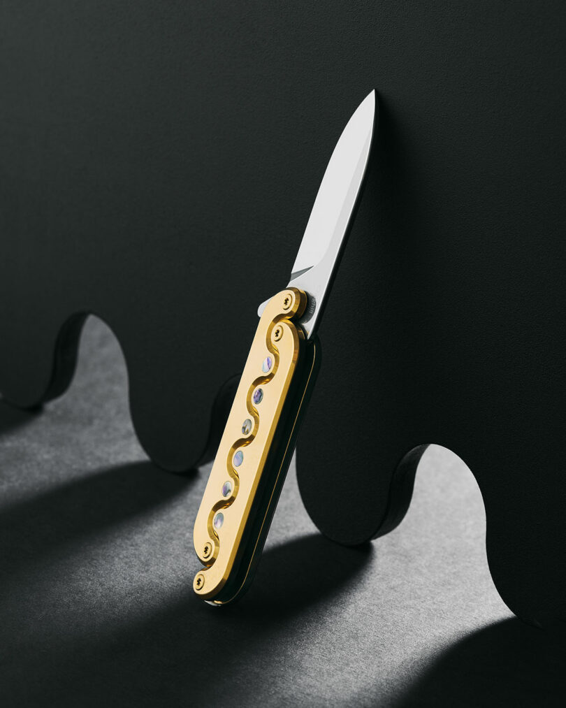 A small pocket knife with a gold handle and intricate design is open and standing upright against a wavy black background.