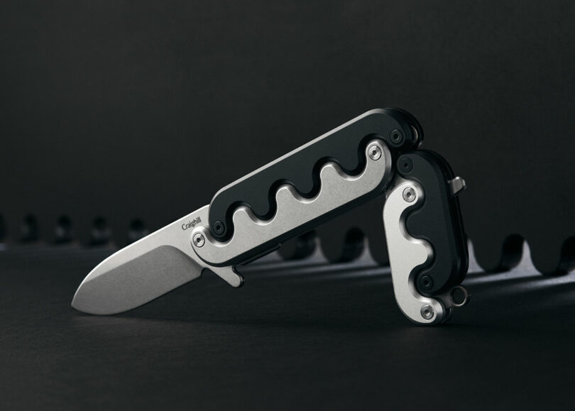 A multi-tool gadget with a partially extended folding knife blade, featuring an intricate metal and black design, displayed against a dark background.