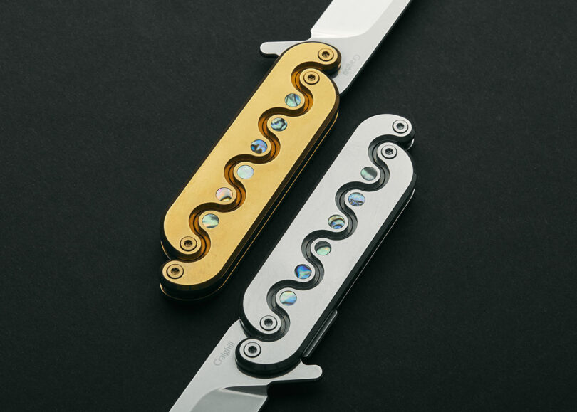 Two folding knives with ornate handles on a black background. One handle is gold and the other is silver, both featuring a wave pattern inlaid with colorful gemstones.