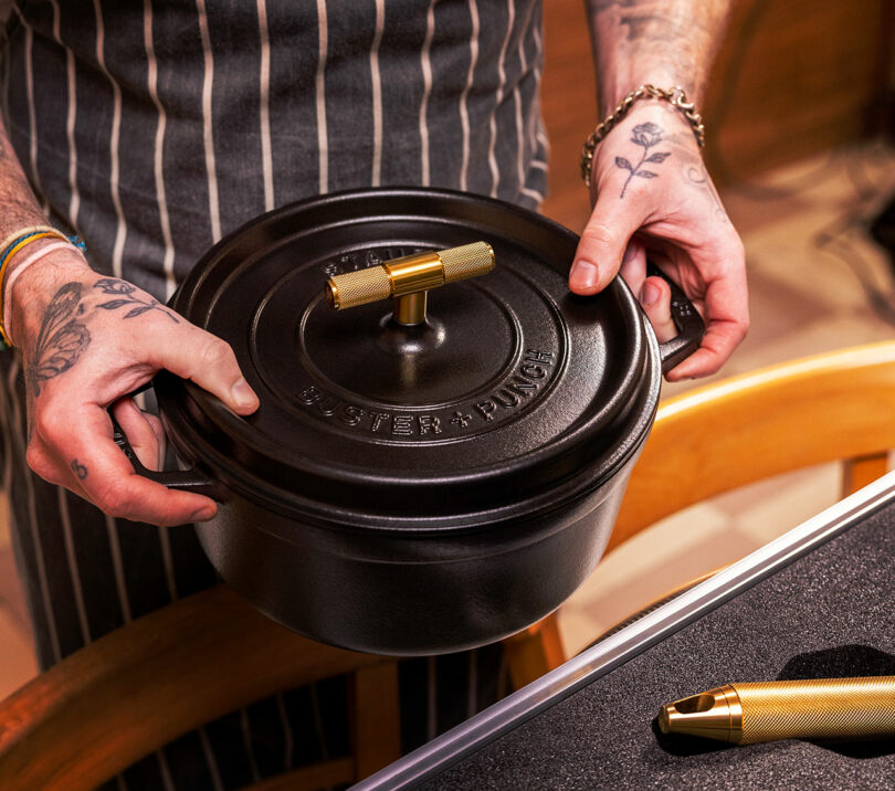 A person wearing a striped apron and bracelets holds a black Staub cast iron pot with a gold handle in both hands. The pot has the words "BUSTER + PUNCH" engraved on the lid.