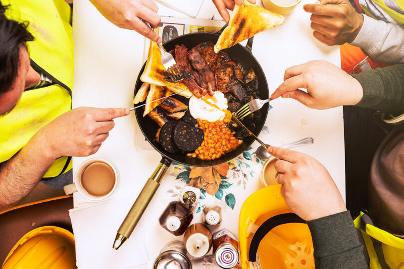 Several people in high-visibility vests reach towards a Staub skillet filled with breakfast items including bacon, beans, and toast, using forks. The table has mugs, condiments, and construction helmets.