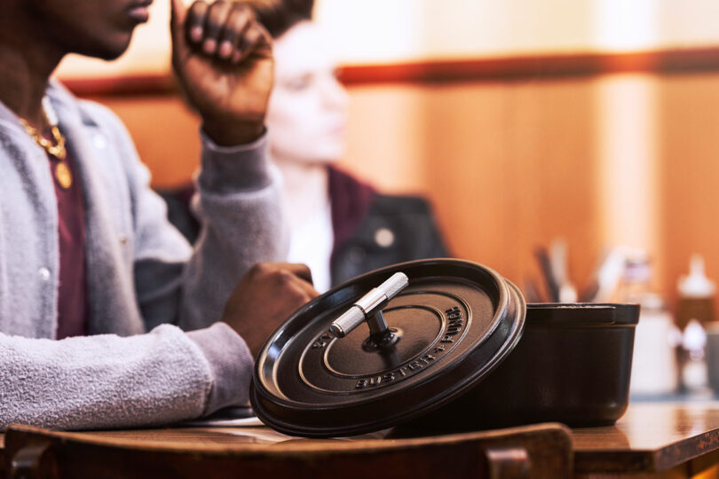 A person sits at a table with a Staub black pot and its lid in the foreground. Another person is seated in the background.