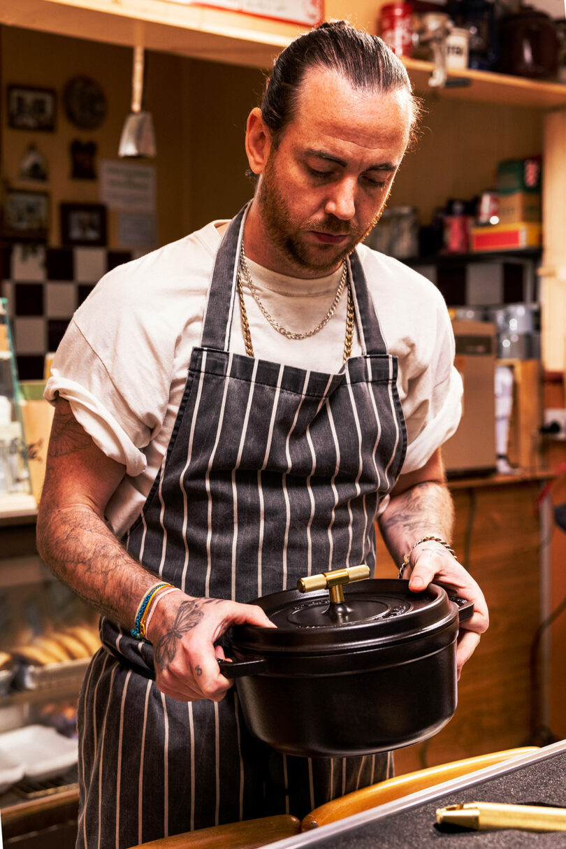 A man wearing a striped apron holds a black Staub cooking pot in a kitchen, with various kitchen items and appliances visible in the background.