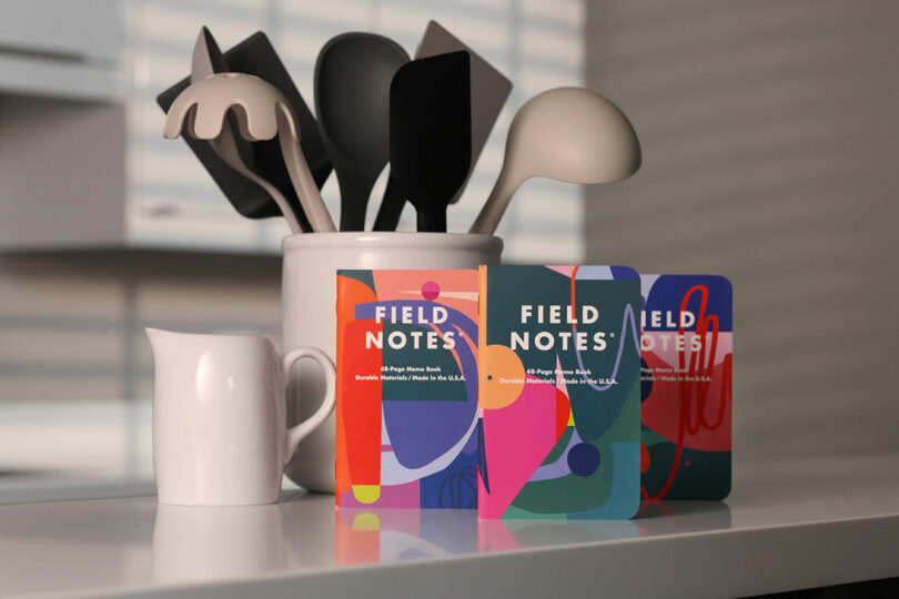 A white countertop displays a white pitcher, a container with black and white kitchen utensils, and three colorful Field Notes notebooks.