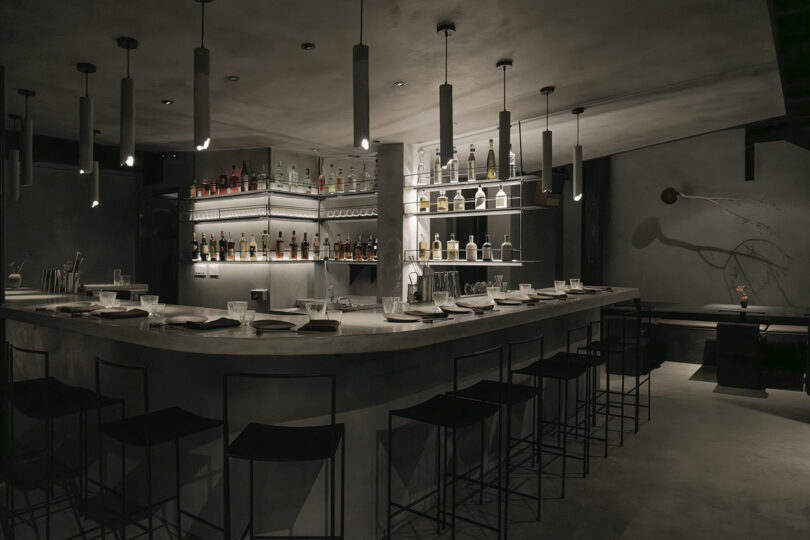 A dimly lit, modern bar with a curved counter and high stools. The bar is stocked with various bottles of liquor, and an overhead light illuminates the area subtly.