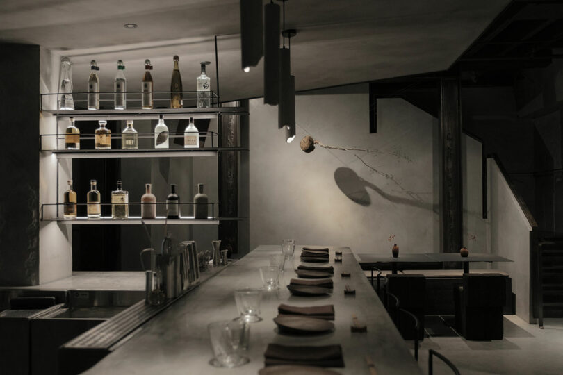 Dimly lit, minimalist bar interior with shelved liquor bottles, a concrete countertop with place settings and glasses, and a hanging light fixture. A sparse decorative branch is on the wall.