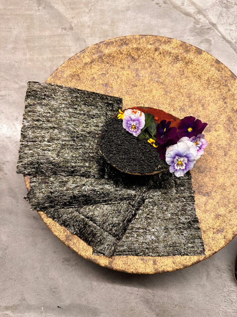 A round dish with seaweed sheets and a small mound of black sesame seeds garnished with purple and white edible flowers on a textured beige plate.