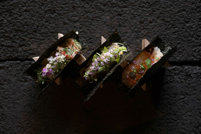 Three gourmet sushi hand rolls, each with unique toppings and garnishes, are displayed in a black holder on a dark textured surface.