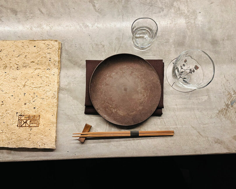 An overhead view of a place setting featuring a round plate, chopsticks, a water glass, and a decorative glass on a textured surface, next to a large textured menu or book.