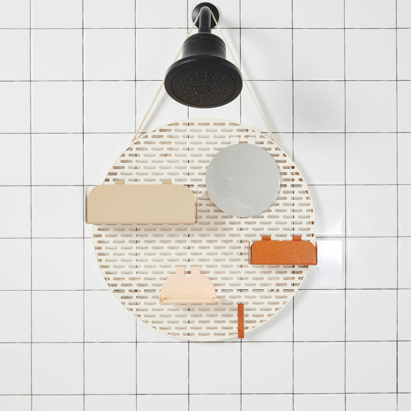 A showerhead above a white oval shower caddy attached to a tiled wall, holding a few beige, grey, and orange items.