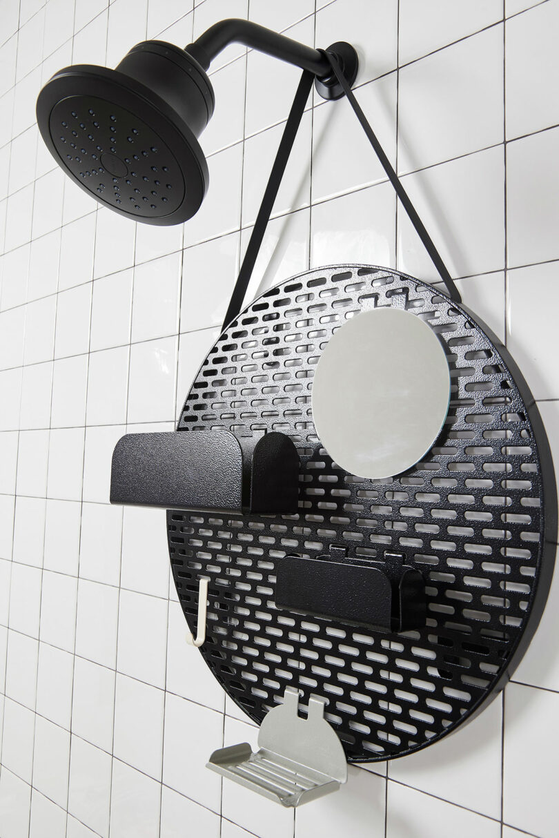 A black showerhead is mounted on a white tiled wall above a black round metal organizer with shelves and a round mirror. The organizer has three shelves and a soap holder.