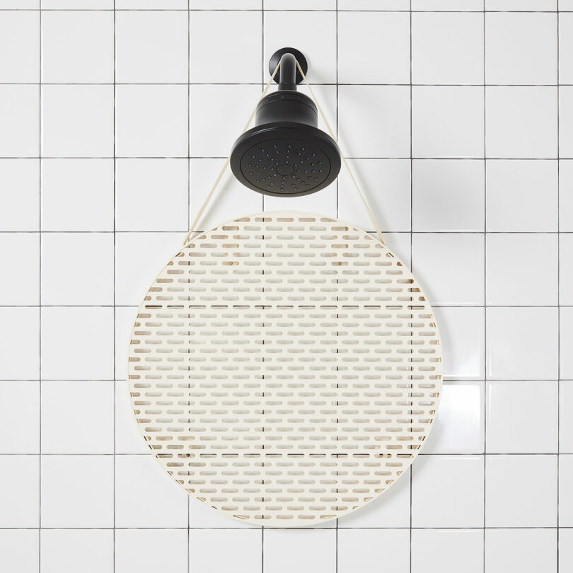 A black showerhead is mounted against white tiled walls, with a round slotted mat hanging below it.