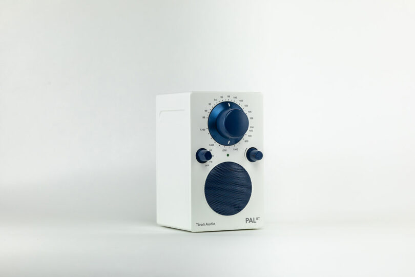 A white Tivoli Audio PAL BT speaker with a circular blue speaker grille, a large blue tuning dial, two smaller blue knobs, and a minimalist design is set against a plain white background.