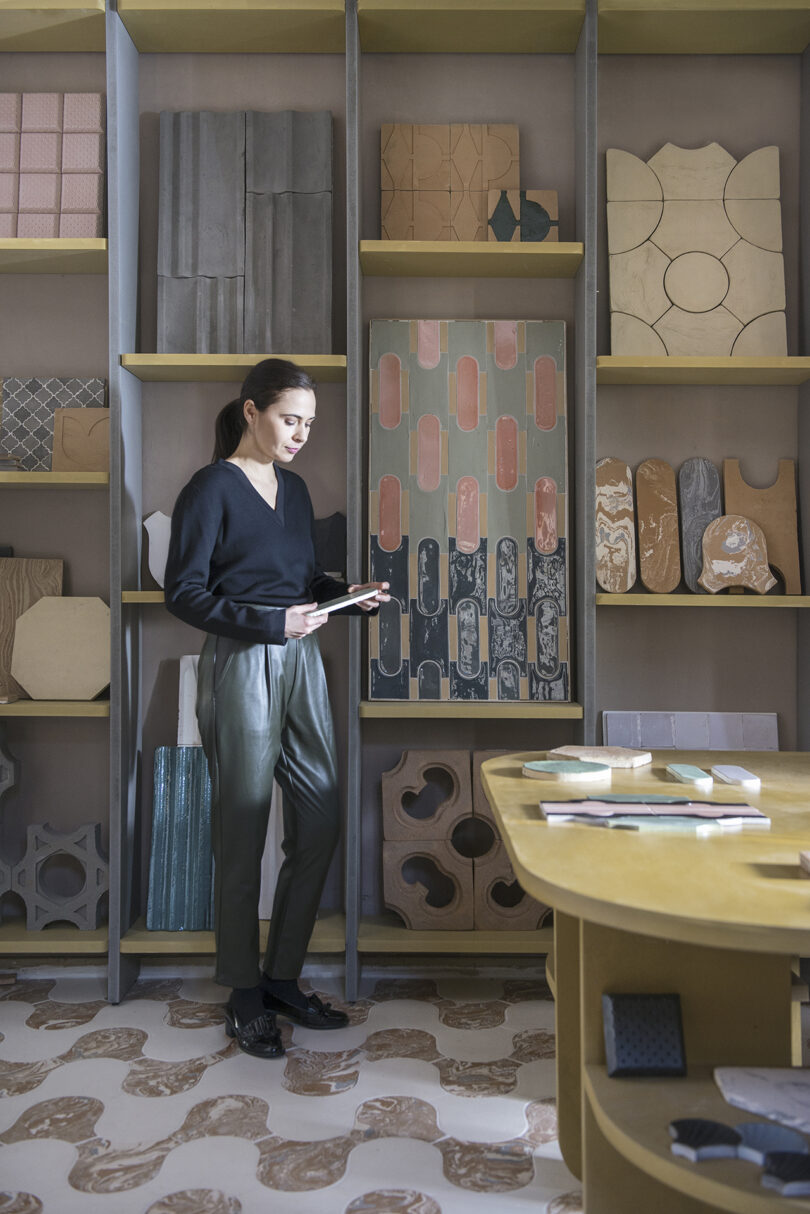 Cristina Celestino stands in a design studio, looking at a tablet. Various tile samples are displayed on shelves behind, and a table with more samples is in the foreground.