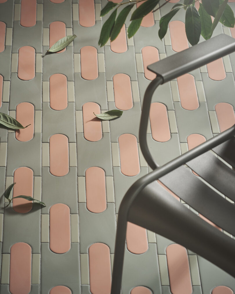 A chair partially visible on a tiled floor with Tivoli Tech green and pink oblong tiles, scattered leaves, and a leafy branch overhead.