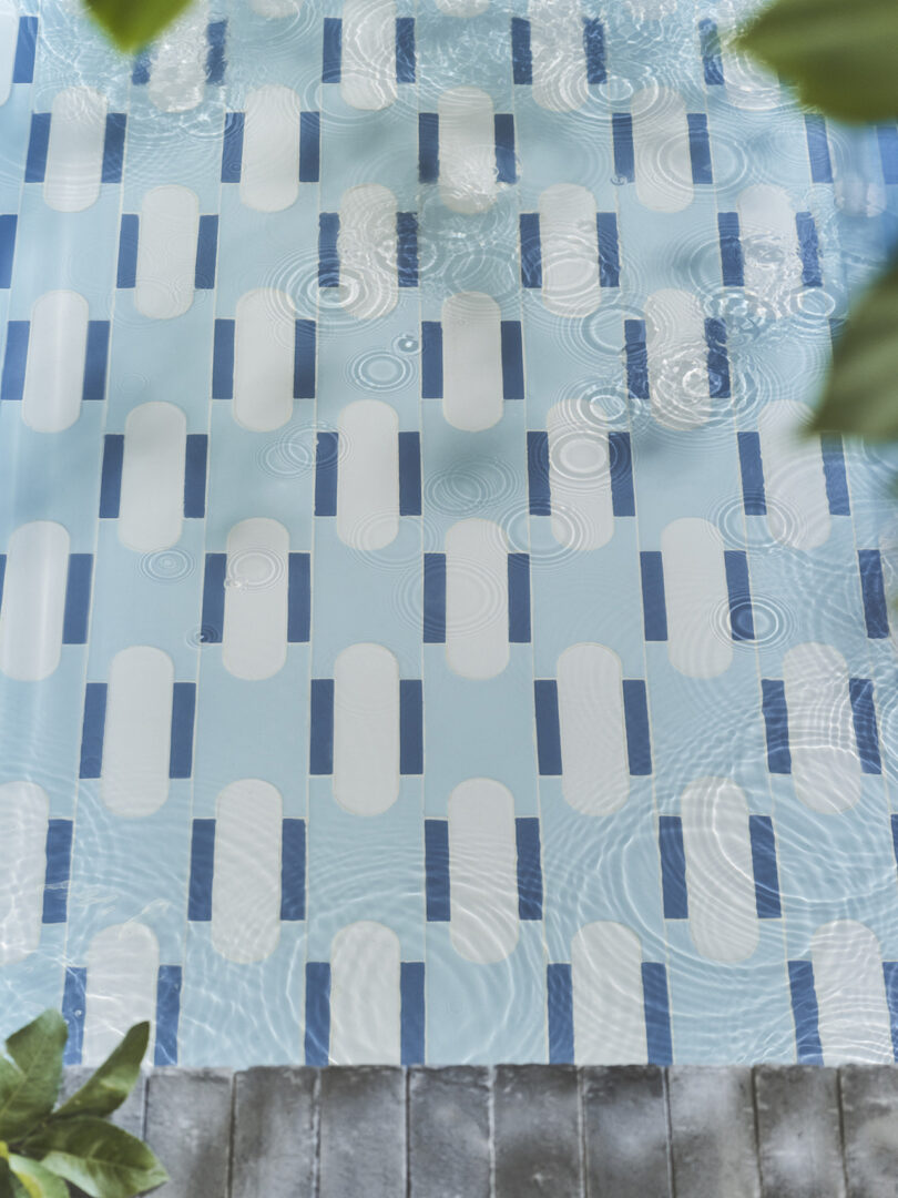 A swimming pool with a blue and white pattern on the floor is shown. The water is rippling, and there are green leaves in the corners of the image.