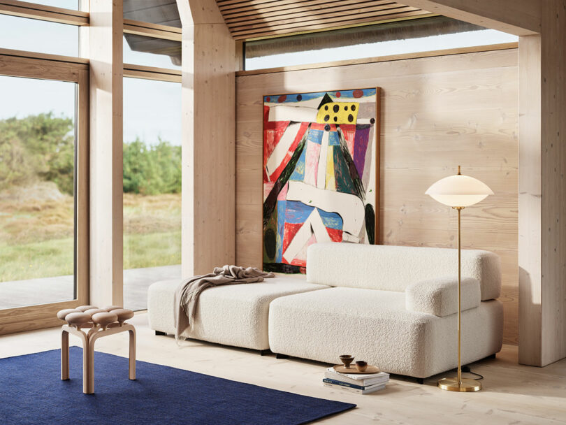 A modern living room featuring a white sectional sofa, a colorful abstract painting, an Utzon Stool, a floor lamp, and large windows. A blue rug covers part of the light wood floor.