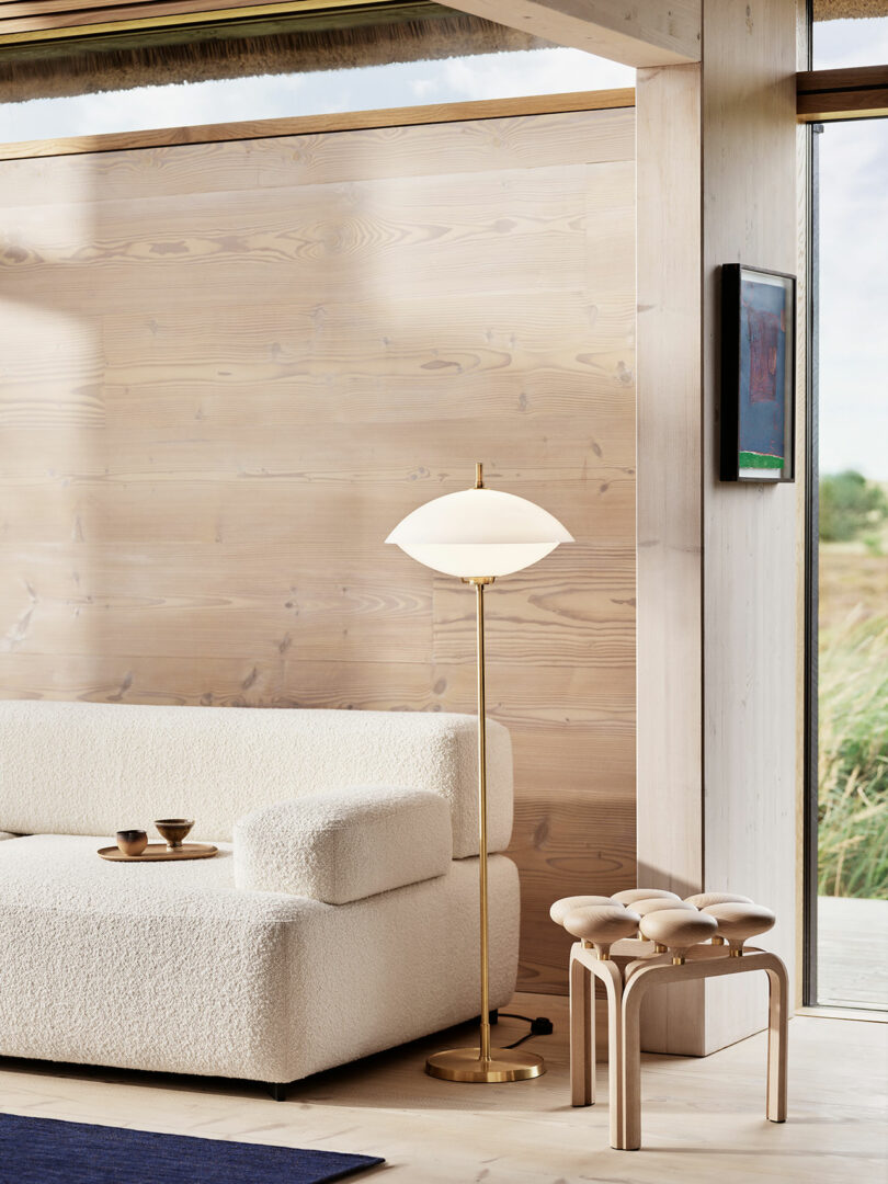 Modern living room with a textured white sofa, a brass floor lamp with a white shade, an Utzon Stool, and a wall-mounted painting. The room features light wood paneling and natural light from a window.