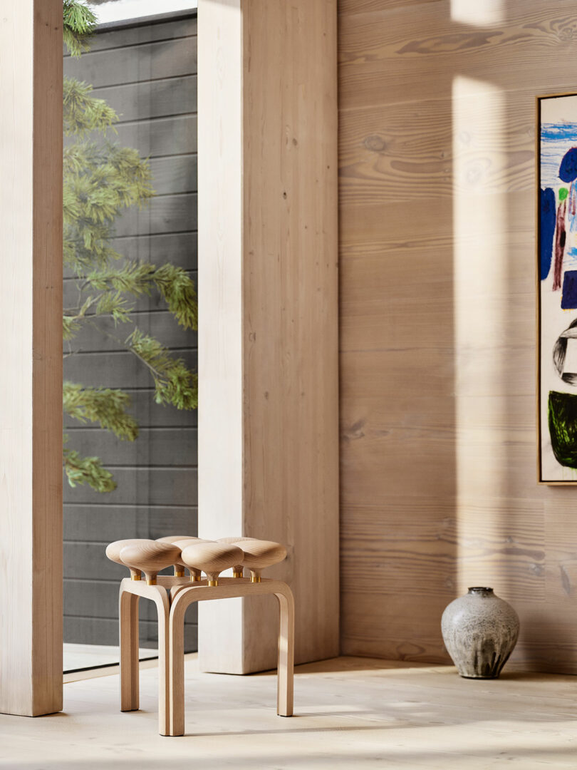 Minimalist interior with an Utzon Stool, ceramic vase, and a modern painting on the wall. Sunlight filters through a window, casting shadows on light wood paneling and greenery visible outside.