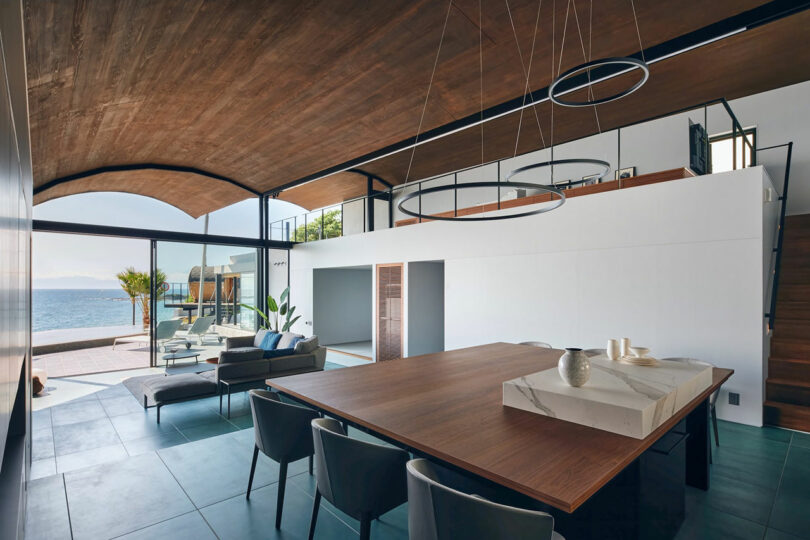 Modern dining and living area with wooden ceiling, large windows, contemporary furniture, and a view of the sea. The space features a dining table with chairs, sofa, indoor plants, and circular light fixtures.