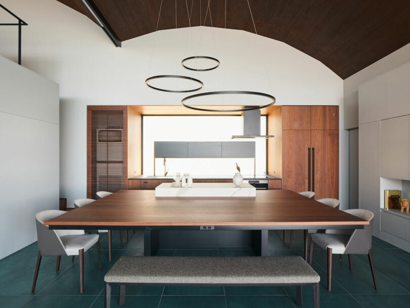 A modern kitchen with a large wooden dining table, six chairs, a bench, circular pendant lights, and built-in wooden cabinetry.