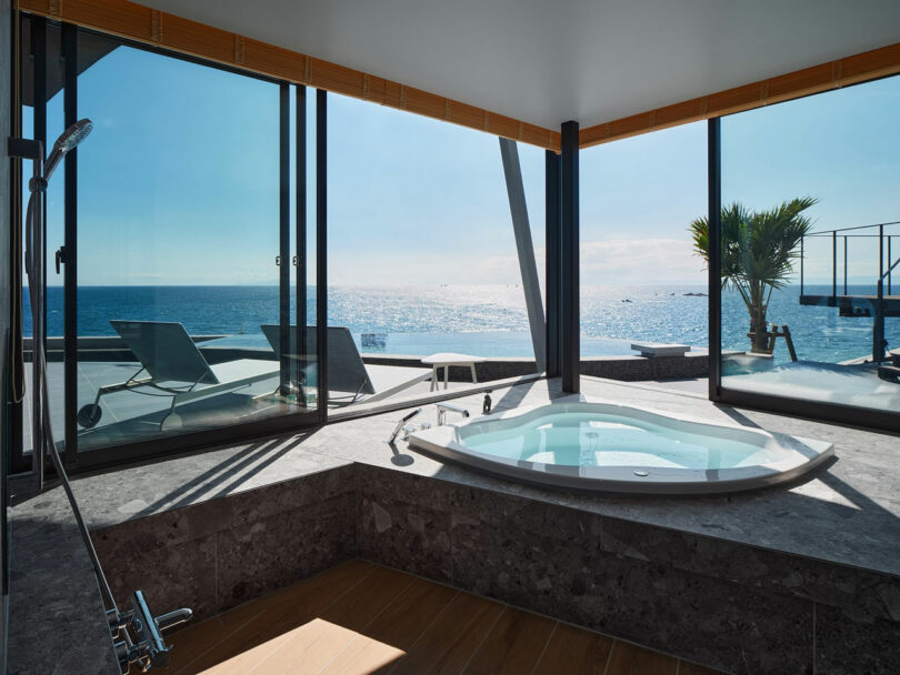 A modern bathroom with a sunken jacuzzi tub, overlooking a sunny ocean view through large glass windows. Two lounge chairs are visible on the patio outside.