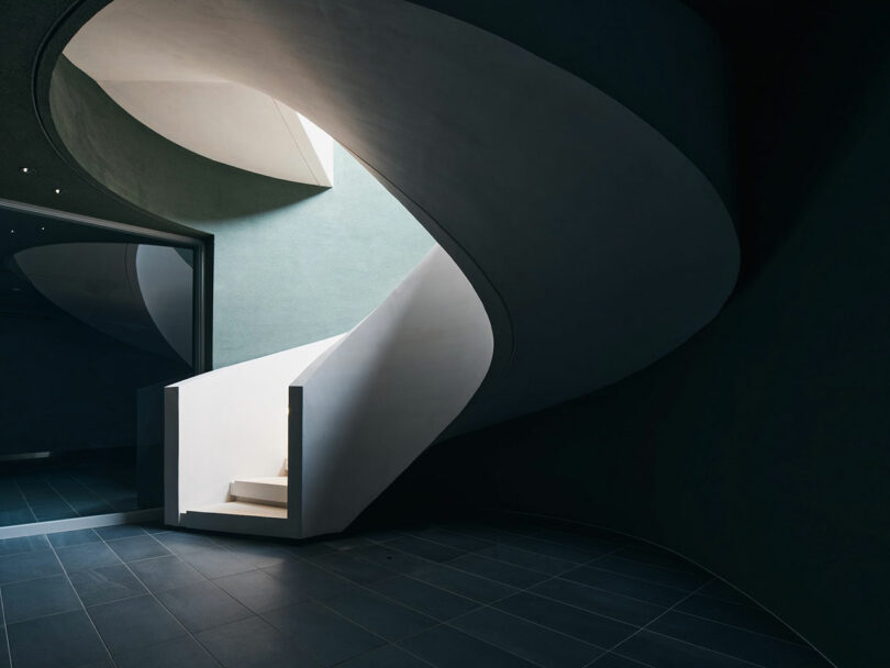 A modern, spiral staircase with sleek white steps and dark walls, illuminated by natural light from a skylight, leads to an upper floor.