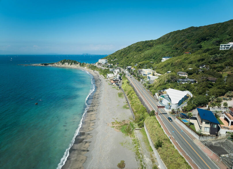 A coastal road runs parallel to a beach with clear blue water and lush green hills in the background, under a clear sky. Sparse buildings and vegetation are visible on the hillside.