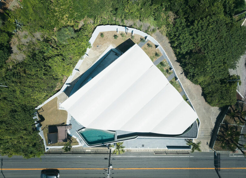 Aerial view of a modern building with a white, multi-peaked roof surrounded by greenery and adjacent to a road.