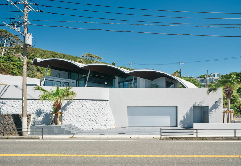 Modern house with a unique wavy roof, white exterior walls, large windows, and palm trees in the front yard, situated along a road with overhead power lines.