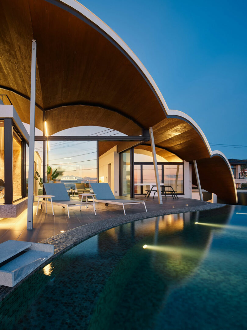 Modern house with a curved wooden roof, large windows, and a pool in the foreground at dusk.