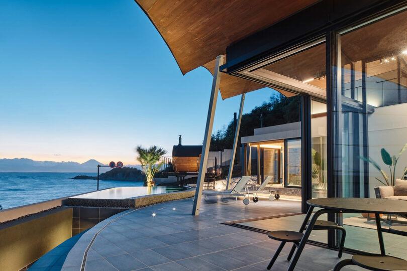 Modern house with large glass windows, an outdoor patio, and an infinity pool overlooking a body of water at dusk. The sky is clear with a hint of twilight.