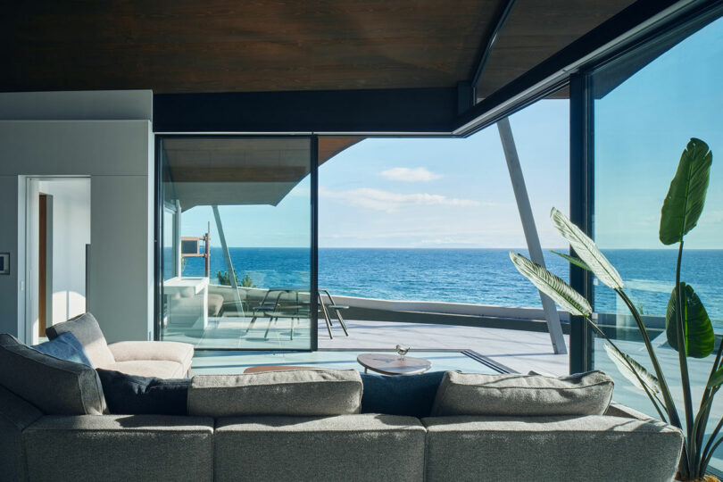 Modern living room with a large gray sectional sofa, large windows, and a view of a balcony overlooking the ocean. A tall plant stands near the window.