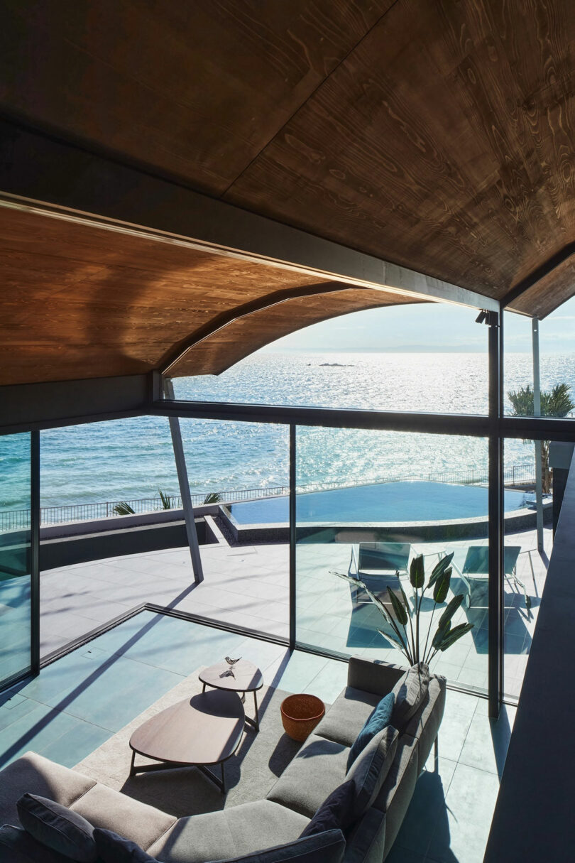 Modern living room with large windows overlooking a sunny seaside view. The room contains a couch, coffee table, and a potted plant, and the patio outside has seating arrangements.