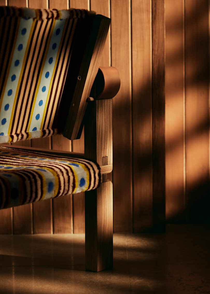 A corner of a wooden chair by Yves Salomon with a colorful, patterned fabric seat and backrest, set against a wooden paneled wall.