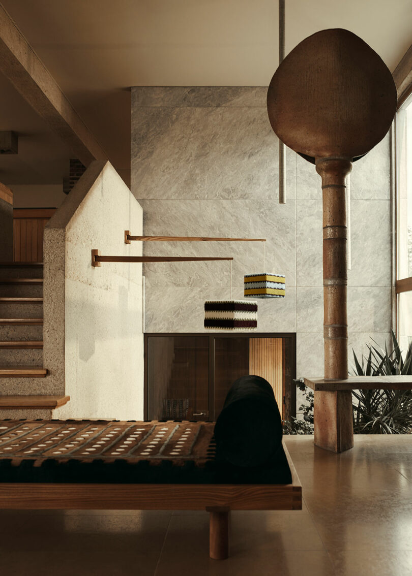 Modern interior with minimalist design featuring a wooden bench, geometric hanging lights by Yves Salomon, large stone pillar, and staircase. The room has neutral tones and large windows allowing natural light in.